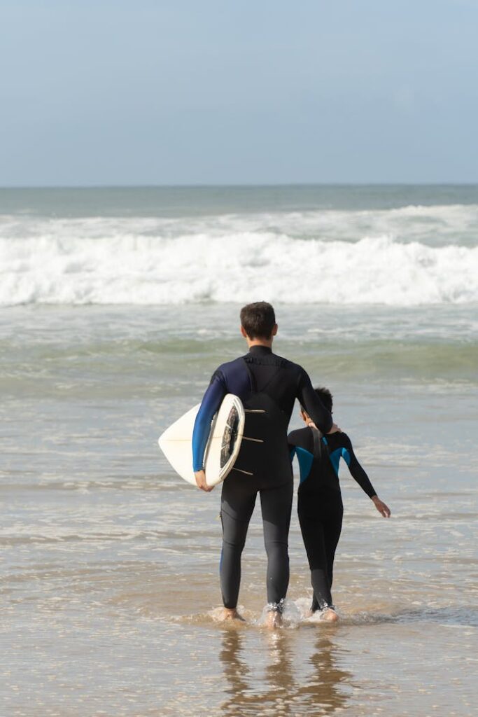 Father and child in wetsuits entering ocean with surfboard in Portugal, enjoying a sunny beach day.